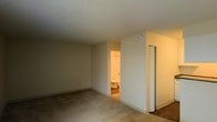 This single-floor residential space presents a clean and functional layout designed for comfortable everyday living. The main room features wall-to-wall carpeting in a neutral tone, contributing to a warm and quiet atmosphere underfoot. The walls are finished in a soft, creamy off-white, lending the space a bright and open feel despite the absence of visible windows in the primary view. A textured popcorn ceiling adds a classic residential character overhead. Accessible directly from the main room is a private bathroom, visible through an open doorway and finished with warm-toned walls and standard fixtures including a toilet, suggesting a full or partial bath. Adjacent to the bathroom entrance is a narrow door, likely leading to a closet, providing convenient built-in storage. The overall design is straightforward and utilitarian, with clean baseboards and simple wall transitions that keep the aesthetic uncluttered. The space is well-suited for use as a bedroom or multipurpose living area, with its en-suite access and storage adding to its practicality. The neutral palette throughout creates a versatile backdrop that accommodates a variety of furnishing styles and personal preferences.