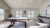 This single-floor entertainment space is defined by its dramatic vaulted ceilings that soar to impressive heights, creating an open and airy atmosphere throughout. The room is flooded with natural light courtesy of an expansive arrangement of windows — including distinctive triangular clerestory windows positioned high along the roofline — that frame lush greenery and mature trees outside. Light wood-style plank flooring runs throughout, complementing the neutral gray-toned walls and bright white trim for a clean, modern aesthetic.

The space is purpose-built for recreation and entertainment, anchored by a full-size pool table with a vivid blue felt surface and a separate air hockey table with a sleek modern base. Wall-mounted cue racks and additional gaming accessories are thoughtfully integrated, keeping the space organized and ready for use. Coastal-themed artwork and decorative greenery add personality and warmth to the otherwise minimal interior.

The layout connects seamlessly to adjacent rooms through wide doorway openings, enhancing the sense of flow and accessibility. Recessed lighting supplements the generous natural light, ensuring the space is well-illuminated at any time of day. The combination of soaring architecture, abundant glazing, and well-curated recreational amenities makes this a standout entertainment destination.
