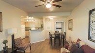 This single-floor unit presents an open-concept layout that seamlessly integrates the living room, dining area, and kitchen into one cohesive space. The living area is outfitted with a ceiling fan with integrated lighting, contributing to both comfort and illumination throughout the space. Warm-toned hardwood-style flooring runs continuously across the main living areas, lending a clean and unified aesthetic.

The kitchen features light wood cabinetry, a white appliance suite including a refrigerator, microwave, and range, as well as dark countertops that provide contrast to the cabinetry. A peninsula counter separates the kitchen from the rest of the open area, offering an informal breakfast bar setup. Track lighting above the kitchen ensures ample task illumination.

The dining area, situated between the kitchen and a hallway, accommodates a four-person table and chairs beneath a chandelier-style light fixture. Adjacent to the dining space, a hallway leads to additional rooms, with what appears to be a washer and dryer unit visible, indicating in-unit laundry. Neutral wall tones throughout contribute to a bright, welcoming ambiance.