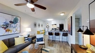 This single-floor space showcases an open-concept layout that integrates the living, dining, and kitchen areas into one cohesive, functional environment. Wood-style plank flooring runs throughout, adding warmth and continuity across the space.

The living area is generously sized, accommodating a full sofa and accent seating, with a ceiling fan providing both comfort and a casual design touch. Neutral grey walls serve as a versatile backdrop, complemented by decorative wall accents that add character without overwhelming the space.

A peninsula-style breakfast bar with dark countertops and barstool seating creates a natural divide between the living area and the kitchen, while maintaining the open feel. The kitchen features white cabinetry, under-cabinet lighting, black appliances, and ample counter space, offering both style and practicality.

Beyond the kitchen, a dining area with a compact table and chairs provides a dedicated space for meals. The overall ambiance is bright and contemporary, with flush-mount lighting and an open layout that maximizes the sense of space throughout.