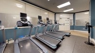 This fitness center offers a comprehensive, single-floor workout facility designed to accommodate a wide range of exercise needs. The space is organized into distinct functional zones, each thoughtfully outfitted with quality equipment. The cardio area features a row of modern treadmills equipped with individual screens, positioned alongside large mirrors that enhance the sense of openness. Adjacent to this, a dedicated strength and free-weight zone includes a full dumbbell rack, stationary bikes, elliptical trainers, and a stability ball area, all set on durable rubberized flooring. A separate strength-training room provides selectorized cable machines, a flat bench, and additional resistance equipment, with bright recessed skylights illuminating the space. The color palette of soft white walls accented by deep teal features gives the facility a clean, modern aesthetic. Framed mirrors throughout the space maximize both light and sightlines. A conveniently located water station with a bottle-fill dispenser is accessible from the main corridor. The facility also includes a fully accessible restroom featuring tile floors and walls, grab bars, a wall-mounted sink with granite countertop, and a vanity mirror, ensuring comfort and convenience for all users.