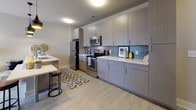 This contemporary single-floor residence offers a thoughtfully designed layout that balances style and functionality throughout. The kitchen serves as a standout feature, showcasing flat-front cabinetry in a warm gray finish, crisp white countertops, and a mosaic tile backsplash that adds a pop of texture and color. Stainless steel appliances, including a range, microwave, and dishwasher, complement the modern aesthetic, while a breakfast bar with seating provides a casual dining option. Light wood-style flooring flows through the main living areas, enhancing the open and airy feel of the space. The living area, visible in the floor plan, provides a comfortable gathering space that flows naturally from the kitchen. The bedroom is a restful retreat, featuring plush carpeting, a ceiling fan with light fixture, and a window dressed with blinds that allows for natural light control. The bathroom is finished with a large vanity mirror, gray wood-tone cabinetry, white countertops, and a full tub-shower combination framed by classic white subway tile. Neutral wall tones and consistent finishes throughout create a cohesive, modern ambiance that is both inviting and easy to personalize.