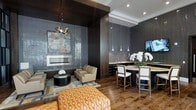 This single-floor space exemplifies contemporary interior design through a carefully curated combination of materials, textures, and lighting. The lounge area anchors the space with a sleek, linear fireplace set against a dramatic textured tile wall in deep charcoal tones, providing a striking focal point. A sculptural chandelier adds artistic flair above a seating arrangement that pairs neutral-toned armchairs with a bold cognac leather tufted ottoman, layered over a geometric area rug atop rich, wide-plank hardwood flooring. A structural dark wood column visually separates the lounge from an adjacent wet bar area, which features dark cabinetry, a light-toned countertop, and a wall-mounted television. Elegant pendant lights hang in a linear arrangement above the bar, casting a warm, refined glow. Bar seating with upholstered white chairs completes the entertaining space, while a bouquet of white orchids adds a natural, sophisticated touch. Recessed lighting throughout enhances the moody ambiance, and the cohesive dark palette, punctuated by warm wood tones and accent pieces, creates a polished, upscale atmosphere ideal for both relaxation and entertaining.