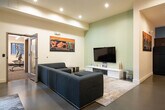 This well-appointed amenity facility is thoughtfully organized into distinct zones, each designed to serve a specific function while maintaining a cohesive contemporary aesthetic throughout.

The lounge area serves as the heart of the space, anchored by a large, deep-seated sectional sofa in charcoal upholstery set atop a patterned area rug. A mounted flat-screen television faces the seating arrangement, while a striking large-format graffiti-inspired artwork commands attention on the wall above, adding an urban, artistic energy to the room. Tall windows along one wall flood the space with natural light and offer views to the exterior.

Adjacent to the lounge is a fully equipped fitness center featuring Precor cardio machines including treadmills, ellipticals, and a stationary bike, as well as a multi-use cable weight machine. Large clerestory windows brighten the gym, and a wall-mounted television and floor-to-ceiling mirror enhance the workout environment.

The kitchenette is finished with rich walnut-toned cabinetry, white countertops, and a teal accent backsplash. It includes a refrigerator, microwave, dishwasher, and sink, providing full utility for gatherings or daily use.

A separate collaborative nook features minimalist tables and mixed seating, set against a raw concrete accent wall and accented with industrial-style lighting, ideal for meetings or focused work.