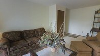 This single-floor space presents a straightforward and functional layout centered around a comfortable living area. The room features neutral-toned walls that create a clean, understated backdrop, contributing to an open and airy feel. The living area is furnished with a patterned sofa in earthy tones, complemented by a glass-topped coffee table adorned with a decorative botanical arrangement that adds a natural, organic touch to the space. Light-colored carpeting covers the floor, adding warmth and texture to the room. A wooden door is visible toward the back of the space, indicating access to an adjacent room or hallway, and a tile or hard flooring transition can be observed near the entryway, suggesting a defined entry zone. The walls are smooth and simply finished, with no pronounced architectural detailing, keeping the aesthetic clean and minimal. The overall ambiance is calm and practical, offering a livable environment with a neutral palette that provides a versatile foundation for personalization. The space is compact yet functional, with a layout that maximizes usable living area on a single level.