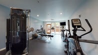 This single-level fitness center is thoughtfully designed to support a full range of workout routines. The space is finished with durable rubber flooring throughout, providing both safety and comfort during exercise. Soft blue-gray walls and recessed lighting create a clean, focused atmosphere conducive to training.

The cardio zone is well-appointed, featuring a treadmill, elliptical trainer, and upright stationary bike, all positioned near large sliding glass doors and windows that flood the area with natural light and offer views of an adjacent outdoor patio. A wall-mounted flat-screen TV is positioned for convenient viewing during cardio sessions.

The strength training area includes a multi-functional cable machine system, a fully stocked dumbbell rack with a wide range of weights, a medicine ball rack with resistance bands, kettlebells, a stability ball, and an adjustable workout bench. These elements together accommodate both free weight and machine-based training.

A full-length mirror is mounted near the entry, supporting form checks during exercise. The space also provides access to a restroom and connects via a corridor to additional amenities. A ceiling-mounted HVAC unit ensures a comfortable environment year-round. Direct access to the outdoor patio further enhances the overall fitness experience.