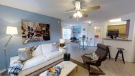 This single-floor space showcases an open-concept layout that seamlessly connects the living and dining areas, creating a versatile and functional environment. The living area is anchored by a white sofa paired with a modern accent chair, centered around a coffee table and complemented by a stylish table lamp. A ceiling fan with integrated lighting fixture serves as both a practical and decorative element overhead.

The walls are painted in a cool blue-gray tone, adding a calm, modern character to the space. Large wall art depicting a classic urban scene adds a bold visual focal point. Light wood-style flooring runs throughout, contributing to a bright and cohesive aesthetic.

Toward the rear, the dining area features a glass-top table with metal folding chairs, positioned near a window that allows natural light to filter into the space. A desk and shelving area are also visible, suggesting a dedicated workspace within the open floor plan. A doorway visible toward the back leads to additional areas of the unit, including what appears to be a kitchen or utility space with shelving.