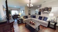 This single-story home offers an inviting open-concept layout that connects the living, dining, and kitchen areas into one cohesive and functional space. The living room serves as the heart of the home, anchored by a generously sized sectional sofa dressed in neutral tones and accented with navy blue throw pillows. A rustic wood coffee table sits at the center, complemented by matching dark wood end tables and stylish table lamps that add warmth to the room. Overhead, an elegant wrought iron chandelier with frosted glass shades provides a refined focal point and ample ambient lighting. Rich hardwood flooring flows throughout, adding depth and continuity across the space. An arched architectural detail frames the transition to the kitchen area, where a raised bar counter and sink are visible, hinting at a practical and well-designed culinary space. The dining area, situated adjacent to the living room, features a dark wood table and chairs alongside a window dressed with flowing curtains that allow natural light to filter in. Decorative wall art and personalized metal lettering lend a homey, personalized character to the walls, completing the warm and welcoming ambiance of this thoughtfully appointed home.