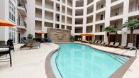 This residential community's amenity and leasing space makes a striking first impression with its soaring double-height lobby, finished in soft grey tones with large-format tile flooring in a crisp two-tone pattern. A dramatic wrought-iron spiral staircase anchors the main lobby, rising to a mezzanine level overlooking the reception area below. The leasing office is thoughtfully appointed with a live-edge wood conference table, mid-century modern chairs, and curated artwork, creating a welcoming yet professional atmosphere. Frosted glass panels, a dedicated workstation, and open display shelving add functional detail throughout the interior space. Large windows and glass-paneled entry doors flood the lobby with natural light while offering direct sightlines to the outdoor courtyard. The exterior courtyard is the centerpiece of the community, featuring a generously sized, freeform swimming pool with vivid turquoise water, surrounded by a wide sun deck lined with chaise lounges and shaded by umbrellas. A stone accent wall with architectural relief detailing and a wood pergola add visual interest to the poolside environment. Multi-story residential buildings with private balconies frame the courtyard, creating an enclosed, resort-like atmosphere. Lush potted palms and decorative planters complete the outdoor setting.