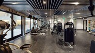 Located at 20 Burling Lane in New Rochelle, NY, this fitness center offers a comprehensive, professionally designed workout facility across a single expansive floor. The space is defined by its striking industrial-modern aesthetic, highlighted by a distinctive slatted metal ceiling with recessed lighting that creates a dynamic, energetic atmosphere throughout.

The main workout floor is generously proportioned, featuring a long row of Spirit treadmills and elliptical machines positioned along floor-to-ceiling windows that invite natural light and views of the surrounding outdoor area. A robust selection of strength-training machines lines the opposite side, complemented by a dedicated free weights section complete with a full dumbbell rack and adjustable benches.

Mirrored glass partitions divide the space elegantly, creating distinct training zones without sacrificing the open feel of the facility. A functional training area includes stability balls and a heavy punching bag, catering to a variety of workout styles. The flooring throughout consists of durable dark rubber gym matting, ideal for high-traffic athletic use.

Private rooms accessible via sleek gray doors provide additional training or utility space. The combination of large windows, polished mirrors, modern equipment, and carefully designed architectural elements makes this fitness center a well-appointed and highly functional destination for all levels of athletic training.