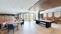 Located in Palm Harbor, Florida, this contemporary amenity lounge is a thoughtfully designed communal space that blends sophisticated aesthetics with versatile functionality. The single-floor layout spans a generous footprint, unified by large-format porcelain tile flooring that flows seamlessly throughout.

The space is organized into distinct yet open zones. A central lounge area anchors the room with plush blue lounge chairs, round pedestal tables, and a curved banquette seating area featuring bold teal and red upholstery beneath vibrant large-scale artwork. Adjacent to this, a dedicated game area showcases a sleek black billiard table set beneath a warm wood slatted ceiling panel, adding texture and depth overhead.

A full kitchen and bar area is equipped with dual islands, wood-accented cabinetry with glass-front upper cabinets, pendant lighting, and white upholstered barstools — ideal for casual gatherings. A built-in booth seating section with slatted wood detailing and archway-shaped backlit wall panels creates an intimate lounge nook with a resort-inspired feel.

Expansive floor-to-ceiling glass doors and windows flood the space with abundant natural light and offer views of the lush Florida outdoors. A dramatic nature-inspired mural adds an artistic focal point, while curated pendant lighting and decorative fixtures enhance the modern, coastal ambiance throughout.