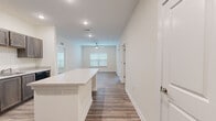 This single-floor residence offers three bedrooms and two bathrooms within a thoughtfully arranged floor plan. The interior is finished with a consistent, contemporary aesthetic characterized by neutral tones, crisp white walls, and wood-look plank flooring that extends throughout the main living areas and bathrooms.

The kitchen serves as a central feature, equipped with dark taupe cabinetry, light stone-look countertops, black appliances, and a generous island with built-in electrical outlets—ideal for both food preparation and casual dining. The kitchen flows directly into the living area, which is brightened by windows fitted with blinds and a ceiling fan overhead.

The primary bedroom is the largest of the three, featuring wood-look flooring, a ceiling fan, and a window for natural light. It connects directly to a private bathroom with a wood-toned vanity, light countertop, and wood-look tile flooring, which in turn leads to a walk-in closet outfitted with wire shelving on multiple walls. The secondary bedrooms are carpeted and served by a second full bathroom with a similar vanity design. Additional closet storage with wire shelving systems provides practical organization throughout the home.