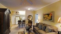 This single-story residence features an open-concept layout that connects the living area and kitchen, creating a cohesive and functional flow throughout the space. The living room is anchored by a comfortable upholstered sofa adorned with decorative pillows, complemented by warm golden-yellow walls that lend an inviting ambiance. A ceiling fan with an integrated light fixture is centrally mounted, enhancing both comfort and illumination. Crown molding adds a refined architectural detail along the ceiling perimeter. An octagonal decorative mirror serves as a focal point on one wall, while framed artwork contributes to the overall warmth of the space. A doorway allows natural light to filter into the room. The kitchen is efficiently designed with white cabinetry, a full-size refrigerator, and a dark countertop that extends into a raised breakfast bar with two bar-height stools, providing casual dining or workspace. The neutral carpeted flooring runs throughout the living area, grounding the warm color palette. Overall, the space presents a cozy, well-appointed interior with thoughtful decorative touches.