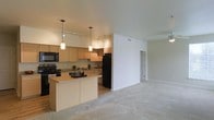This single-floor residence showcases an open-concept layout that connects the kitchen and living area in a cohesive, functional design. The kitchen is equipped with black appliances, including a refrigerator, range, and over-the-range microwave, complemented by light wood cabinetry that lines the walls and provides ample storage. A peninsula with a light countertop extends into the space, offering additional prep area and serving as a natural divider between the kitchen and living room. Two pendant lights hang above the peninsula, while a track lighting system illuminates the kitchen workspace, creating a warm, well-lit environment. The flooring transitions from hard surface in the kitchen to plush, light-toned carpet in the living area, adding comfort underfoot. The living space is generously proportioned, featuring neutral walls and a clean, open feel that allows for flexible furniture arrangement. A hallway visible in the background suggests access to additional rooms within the unit. The overall aesthetic is contemporary and understated, with consistent neutral tones throughout that create a calm, move-in-ready atmosphere.