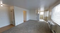 This single-floor space features an open-concept layout that seamlessly connects the living area to the kitchen. The living area is finished with wall-to-wall carpet in a neutral tone, contributing to a comfortable and understated atmosphere. White walls throughout the space lend a clean, bright feel, while the flat ceiling with a flush-mounted light fixture keeps the design simple and functional.

A distinctive built-in open shelving unit serves as a visual divider between the living area and the kitchen, offering both storage and an architectural detail that adds character to the otherwise straightforward layout. The kitchen is finished with tile flooring, providing a practical contrast to the carpeted living space. A white refrigerator is visible, and a window in the kitchen area allows natural light to filter into the space, brightening the rear of the unit.

A doorway off the living area leads to an additional room or hallway, suggesting further functional space beyond what is immediately visible. Overall, the unit presents a practical, move-in-ready layout with a neutral palette suitable for a variety of furnishing styles.