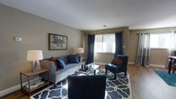 This single-floor residence offers a thoughtfully designed interior that balances comfort and style. The living room serves as the heart of the home, featuring warm hardwood flooring, neutral taupe walls, and large windows dressed with floor-length navy drapes that allow abundant natural light to fill the space. A plush gray sofa anchors the seating area, complemented by teal and rust accent pillows, a glass-top coffee table, and a bold geometric patterned area rug in navy and white. A teal accent chair adds a pop of color, while framed wall art and coordinating table lamps contribute to the cohesive, curated aesthetic. The dining area flows naturally from the living space, centered around a dark wood dining table surrounded by rich brown leather chairs. A pendant light fixture with a frosted glass bowl shade hangs overhead, casting warm, focused light across the table. Hardwood flooring continues throughout, and a vibrant teal area rug beneath the dining set adds a lively contrast. A colorful abstract wall painting provides an artistic focal point. Together, these spaces create a warm, inviting atmosphere ideal for both everyday living and entertaining.
