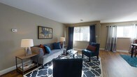 This single-floor residence offers a thoughtfully designed interior that balances comfort and style. The living room serves as the heart of the home, featuring warm hardwood flooring, neutral taupe walls, and large windows dressed with floor-length navy drapes that allow abundant natural light to fill the space. A plush gray sofa anchors the seating area, complemented by teal and rust accent pillows, a glass-top coffee table, and a bold geometric patterned area rug in navy and white. A teal accent chair adds a pop of color, while framed wall art and coordinating table lamps contribute to the cohesive, curated aesthetic. The dining area flows naturally from the living space, centered around a dark wood dining table surrounded by rich brown leather chairs. A pendant light fixture with a frosted glass bowl shade hangs overhead, casting warm, focused light across the table. Hardwood flooring continues throughout, and a vibrant teal area rug beneath the dining set adds a lively contrast. A colorful abstract wall painting provides an artistic focal point. Together, these spaces create a warm, inviting atmosphere ideal for both everyday living and entertaining.