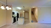 This two-story home offers a well-designed open-concept main level that seamlessly connects the kitchen and living areas. The kitchen is equipped with white cabinetry, dark countertops, and black appliances, including a refrigerator, stove, and built-in microwave. A central island with a sink and pendant lighting above serves as both a functional workspace and a natural dividing point between the kitchen and the adjacent living area. Recessed lighting and a flush ceiling fixture provide ample illumination throughout. Crown molding adds a polished architectural detail along the ceiling line. The main level features neutral-toned carpeting that extends throughout the living space, contributing to a warm and cohesive feel. A door near the base of the staircase leads to an entryway or secondary access point. Carpeted stairs with a wooden handrail and metal balusters lead to the upper floor, which provides additional living or sleeping quarters. The overall color palette—warm beige walls, white trim, and dark kitchen accents—creates a balanced and inviting atmosphere throughout the home.
