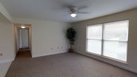 This single-story space presents a practical and welcoming layout designed for comfortable everyday living. The main living area is finished with wall-to-wall carpeting in a neutral beige tone, providing a warm and soft underfoot feel throughout the room. Smooth, light-colored walls create a clean and airy atmosphere, complemented by crisp white baseboards that add a polished finish to the space. A ceiling fan with a built-in light fixture is centrally mounted overhead, offering both ambient lighting and air circulation year-round. Natural light flows generously into the room through a large window fitted with horizontal blinds, allowing for adjustable light control and privacy. Multiple electrical outlets are conveniently distributed along the walls, reflecting thoughtful and functional design. The open feel of the room provides flexibility for a variety of furniture arrangements and uses. The overall ambiance is calm and neutral, making it a versatile space that can be easily personalized to suit a range of preferences and lifestyles.