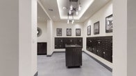This mail and package room is designed with both functionality and aesthetic cohesion in mind. The space is finished with large-format gray tile flooring that extends throughout, lending a clean and contemporary feel. Neutral, warm-toned walls provide a bright backdrop, complemented by recessed lighting and a decorative tray ceiling with a multi-arm pendant fixture that serves as a subtle focal point.

Banks of dark-finished individual mailboxes line the walls, offering organized mail retrieval for a large number of residents. A central island with a dark wood-grain cabinet base and a stone-style countertop provides a convenient surface for sorting mail, complete with a built-in stainless steel sink. Additional dark cabinetry is positioned along the perimeter walls for supplementary storage.

A dedicated package locker system is integrated into one section of the room, allowing secure retrieval of parcels at any time. Framed black-and-white postal stamp artwork is displayed above the mailbox banks, reinforcing the room's cohesive postal theme. An adjacent corridor with patterned carpet connects the space to other areas of the building. Overall, the room balances practical postal infrastructure with polished, modern design details.