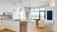 This thoughtfully designed single-floor unit showcases a contemporary aesthetic defined by clean lines, an open layout, and an abundance of natural light. The open-concept living and kitchen area forms the heart of the space, where exposed concrete ceiling panels create an industrial contrast against crisp white walls and warm honey-toned hardwood flooring. Floor-to-ceiling black-framed windows frame sweeping city skyline views, flooding the living area with daylight and providing a striking architectural focal point.

The kitchen is outfitted with high-gloss white cabinetry extending to the ceiling, a suite of stainless steel appliances including a full-sized refrigerator, range, microwave, and dishwasher, and a generous island with a light quartz countertop and pendant lighting overhead. Open shelving adds functional display space near the living area transition.

The bedroom continues the unit's design language, featuring the same exposed concrete ceiling detail, hardwood floors, and a large black-framed window with city views, creating a calm, airy retreat.

The bathroom offers a refined, spa-like finish with light wood vanity cabinetry, a undermount sink with brushed gold fixtures, large-format neutral tile, and a frameless glass walk-in shower — completing a cohesive, modern living experience throughout.