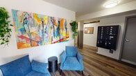 Located in the University District neighborhood of Seattle, Washington, this building's common areas offer a thoughtfully designed environment for residents. The entry lobby sets a welcoming tone with warm wood-look plank flooring, crisp white walls, and an abundance of natural light streaming through large windows and a glass entry door. A comfortable seating area furnished with plush blue armchairs invites residents to relax, while a bold, large-scale abstract painting featuring vivid yellows, blues, and reds serves as a striking focal point. Lush trailing greenery adds a natural, refreshing touch to the space. Practical amenities are seamlessly integrated into the lobby, including a multi-unit mailbox console and a building intercom and fire safety panel mounted on the wall. Track lighting overhead ensures the space is well-lit at all hours. Extending from the lobby, a corridor with the same wood-look flooring leads to a dedicated bike storage and utility area. This lower-level space features wall-mounted and floor bike racks, a communal high-top table with seating, exposed overhead utility pipes that lend an industrial character, and additional colorful artwork that keeps the aesthetic cohesive and lively throughout the building's shared spaces.