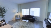 This single-floor space presents a thoughtfully designed living area that balances modern aesthetics with functional comfort. The living room is defined by its clean architectural lines, neutral wall tones, and wood-style flooring that runs throughout, creating a cohesive and inviting atmosphere.

Two generously sized windows fitted with horizontal blinds allow abundant natural light to fill the room, offering views of the surrounding urban environment. The bright, airy quality of the space is further enhanced by the notable ceiling height, which contributes to an open and unenclosed feel.

The living area is well-appointed, featuring a dark upholstered sofa accented with decorative pillows, paired with a coordinating accent chair draped with a casual throw. A geometric-patterned area rug anchors the seating arrangement, while a wire-frame coffee table and matching side table add contemporary detail without visual weight. A sleek arc floor lamp provides both functional and ambient lighting, completing the modern, urban character of the space. The overall design is minimal yet welcoming, suited for comfortable everyday living.
