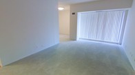 This single-floor residence offers a clean, simple layout designed for comfortable everyday living. The main living area is finished with wall-to-wall carpeting in a soft, neutral tone that contributes to a calm and understated ambiance throughout the space. Bright white walls keep the interior feeling open and airy, providing a versatile backdrop suited to a variety of décor styles.

A large window fitted with vertical blinds serves as a prominent feature of the living area, allowing abundant natural light to filter in and brighten the room. The blinds offer flexible light control, making it easy to adjust the atmosphere as needed throughout the day.

A centrally positioned ceiling light fixture provides consistent overhead illumination, ensuring the space remains well-lit regardless of the time of day. A ceiling-mounted HVAC vent indicates a central air system, contributing to year-round climate comfort. An adjacent room, visible through an open passage, mirrors the same carpeted flooring and neutral palette, suggesting a cohesive design language carried throughout the home.

Overall, the space presents a tidy, move-in-ready environment with a straightforward and functional layout, ideal for those seeking a clean and low-maintenance living space on a single level.