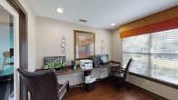 This business center is a well-appointed, single-floor space designed to meet the productivity needs of its users. The room features rich hardwood flooring throughout, lending a warm and professional ambiance that is complemented by neutral-toned walls and recessed lighting overhead.

The centerpiece of the space is a continuous built-in wooden workstation that spans one wall, accommodating multiple desktop computers, monitors, keyboards, and a multifunction printer, making it well-suited for a range of tasks from document printing to general computing. Ergonomic executive chairs in brown leather provide comfortable seating at each station.

Decorative touches elevate the functional setting — a large gold-framed piece of colorful artwork anchors the main wall, flanked by modern circular metal wall accents. Vibrant orange and patterned fabric valances frame the windows, adding a pop of color against the white horizontal blinds that allow for natural light control. Multiple windows offer views of the surrounding greenery outside.

A floating shelf along another wall serves as an information station, topped with brochures and a cork bulletin board displaying community notices and resources. A doorway connects the business center to an adjacent tiled corridor, providing easy access to the broader facility. The overall design balances practicality with a welcoming, polished aesthetic.