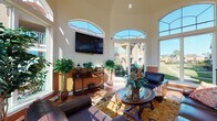 This single-floor commercial space exudes a warm, upscale ambiance blending elegant design with functional areas suited for hospitality or professional use. The centerpiece is a grand lounge area distinguished by soaring ceilings and a striking arrangement of tall arched windows that flood the room with abundant natural light and offer open views of lush green surroundings. Rich hardwood flooring, ornate area rugs, and leather seating arrangements create a refined yet inviting atmosphere, complemented by a wall-mounted flat-screen television and classic wooden console furniture.

Adjacent to the lounge, a dedicated workspace features a substantial wooden desk with dual monitors, positioned beside large glass doors that extend the sense of openness to the outdoors. The dining area is appointed with a round glass-top table, upholstered leather chairs, and a decorative wooden sideboard with a granite top, all set atop light travertine tile flooring that continues throughout the transitional spaces.

A professional front reception desk anchors the administrative area, outfitted with workstations and framed by plantation-shuttered French doors. Nearby, the charming "Cafe Bar Amalfi" station offers a coffee service nook with large windows lining one wall, adding a bright, welcoming touch. Two-tone warm beige and terracotta wall treatments tie the entire space together cohesively.