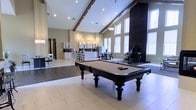 This impressive single-floor amenity space is defined by its soaring vaulted ceilings with exposed beams, creating an open and airy atmosphere that immediately commands attention. Large, floor-to-ceiling windows flood the interior with generous natural light, enhancing the bright and welcoming ambiance throughout. The expansive main area serves as the centerpiece of the space, anchored by a stately billiards table with ornate carved legs, positioned atop light-tiled flooring that contrasts beautifully with the darker flooring zones nearby. A dramatic dark accent column adds a bold architectural element, framing a grand piano that rests in a dedicated corner — an elegant touch that lends a sophisticated, upscale character to the room. Recessed lighting and pendant fixtures provide warm, layered illumination throughout. Toward the rear, a fully equipped kitchen features white cabinetry with glass-front upper cabinets, a full-size refrigerator, countertop appliances, and bar stool seating — ideal for hosting gatherings. A casual dining area with table and chair seating sits adjacent, completing the entertainment-ready layout. The overall design seamlessly blends modern elegance with functional amenities, making this a versatile and inviting communal space.