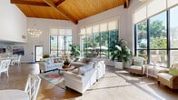 Located at 300 W Airport Blvd in Sanford, Florida, this expansive single-floor community space offers a welcoming and airy atmosphere across a generously sized open floor plan. The architectural design is immediately striking, highlighted by a dramatic vaulted ceiling with warm, exposed wood beams and tongue-and-groove wood paneling that anchors the main living area with natural warmth and character. Light-toned herringbone-patterned hardwood flooring flows seamlessly throughout, lending a sophisticated yet approachable aesthetic.

The living area is bathed in abundant natural light, courtesy of floor-to-ceiling gridded black-frame windows that frame lush outdoor greenery and views of the surrounding landscape. Elegant chandelier fixtures and recessed lighting provide layered illumination throughout the day and evening. The seating area is furnished with plush white sofas and accent chairs arranged around a central coffee table, complemented by vibrant tropical botanical rugs and lush indoor plants that reinforce a Florida-inspired ambiance.

A dining area with crisp white round tables and cross-back chairs transitions smoothly into additional lounge seating, including floral accent chairs positioned near a wall-mounted television and media console. Colorful artwork and botanical paintings adorn the bright white walls throughout, contributing to a lively yet refined atmosphere. A service counter and additional functional areas are visible toward the rear of the space, enhancing its versatility as a communal gathering destination.
