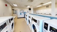 Located at 808 Memorial Dr in Cambridge, MA, this shared laundry facility is a well-equipped, single-floor space designed to efficiently serve its users. The room is outfitted with a generous row of front-loading washing machines along one side, complemented by a bank of top-loading dryers along the opposite wall, providing ample capacity for simultaneous loads. A built-in folding table offers a dedicated surface for organizing clean laundry, while instructional signage for proper washing and drying techniques is posted on the walls for user reference. A centrally located payment kiosk and an information bulletin board make the facility easy to navigate and manage. The space features a drop-off laundry bin near the entrance, offering added convenience. Seating is available along the windowed wall, where large windows flood the room with natural light and offer views of the surrounding Cambridge streetscape. The interior maintains a clean, functional aesthetic with light-colored walls accented by soft blue tones, durable flooring, and a drop ceiling with recessed fluorescent lighting. The overall layout is open and practical, prioritizing ease of use and comfort for all visitors.