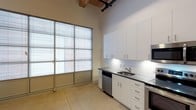 Unit 137 is a single-floor space that masterfully blends industrial character with modern finishes. The interior immediately draws attention with its striking exposed timber columns and warm wood-beam ceiling, which run throughout and lend a sense of historic authenticity. Black exposed utility pipes trace the ceiling, reinforcing the raw, industrial aesthetic while adding architectural interest.

The polished concrete flooring extends throughout the space, providing a durable and visually cohesive foundation that complements the warm tones of the wood overhead. A long corridor flanked by crisp white walls connects the various areas of the unit, with a white paneled door offering access to additional spaces.

The kitchenette is a standout feature, equipped with sleek white cabinetry, dark countertops, a stainless steel sink, a dishwasher, and additional stainless steel appliances. A white subway tile backsplash adds a clean, contemporary touch. Large industrial-style windows fitted with horizontal blinds flood the kitchen area with natural light and offer views of the surrounding building exterior, creating a bright and functional workspace.

Overall, the space strikes a compelling balance between preserved industrial elements and thoughtfully updated modern amenities, making it a versatile and visually distinctive environment.