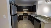 This single-floor space is thoughtfully laid out to maximize functionality and comfort across its key living areas. The kitchen stands out as a highlight of the unit, showcasing dark espresso cabinetry that extends from floor to ceiling, complemented by granite countertops with subtle speckled detailing. Stainless steel appliances — including a gas range, over-the-range microwave, and a full-size refrigerator — provide a sleek, cohesive aesthetic. Light wood-tone flooring in the kitchen adds warmth and contrast against the bold cabinetry, while an undermount sink is neatly integrated into the countertop layout.

The living area is generously proportioned, offering ample room for seating and furnishings. Natural light filters in through windows positioned along the exterior wall, brightening the neutral-toned interior. A dedicated sleeping area provides a private retreat, and additional spaces — including what appears to be a bathroom and entryway — complete the unit's practical floor plan.

Throughout the space, the design balances modern finishes with a functional layout. Neutral wall tones and consistent flooring materials create a unified and inviting atmosphere, making this a well-rounded space suited for everyday living.