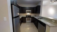 This single-floor space is thoughtfully laid out to maximize functionality and comfort across its key living areas. The kitchen stands out as a highlight of the unit, showcasing dark espresso cabinetry that extends from floor to ceiling, complemented by granite countertops with subtle speckled detailing. Stainless steel appliances — including a gas range, over-the-range microwave, and a full-size refrigerator — provide a sleek, cohesive aesthetic. Light wood-tone flooring in the kitchen adds warmth and contrast against the bold cabinetry, while an undermount sink is neatly integrated into the countertop layout.

The living area is generously proportioned, offering ample room for seating and furnishings. Natural light filters in through windows positioned along the exterior wall, brightening the neutral-toned interior. A dedicated sleeping area provides a private retreat, and additional spaces — including what appears to be a bathroom and entryway — complete the unit's practical floor plan.

Throughout the space, the design balances modern finishes with a functional layout. Neutral wall tones and consistent flooring materials create a unified and inviting atmosphere, making this a well-rounded space suited for everyday living.