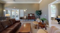 This single-floor community clubhouse offers a thoughtfully designed gathering space with a warm, welcoming ambiance throughout. The main lounge serves as the centerpiece of the facility, featuring a striking stone fireplace framed by elegant white columns and crown molding. Comfortable leather seating and classic wood furnishings create an inviting atmosphere, while arched windows and French doors allow generous natural light to fill the room.

Adjacent to the lounge is a dedicated game room, highlighted by a full-size billiards table positioned beneath a pendant light fixture. Tall arched windows overlook lush outdoor greenery, and a wall-mounted television adds to the recreational appeal. A separate business center, enclosed by glass-paneled French doors, provides multiple computer workstations for residents.

The fully equipped kitchen features rich wood cabinetry, dark granite countertops, tile flooring, and a full suite of appliances including a refrigerator and microwave, making it ideal for hosting community events. Near the entrance, a cozy bistro nook with a high-top table and bar seating, accented by stone tile flooring and transom windows, provides a welcoming transition into the space. Throughout, earthy taupe tones, carpet flooring, and tasteful décor create a cohesive and comfortable community environment.