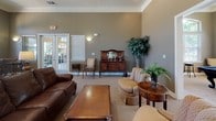 This single-floor community clubhouse offers a thoughtfully designed gathering space with a warm, welcoming ambiance throughout. The main lounge serves as the centerpiece of the facility, featuring a striking stone fireplace framed by elegant white columns and crown molding. Comfortable leather seating and classic wood furnishings create an inviting atmosphere, while arched windows and French doors allow generous natural light to fill the room.

Adjacent to the lounge is a dedicated game room, highlighted by a full-size billiards table positioned beneath a pendant light fixture. Tall arched windows overlook lush outdoor greenery, and a wall-mounted television adds to the recreational appeal. A separate business center, enclosed by glass-paneled French doors, provides multiple computer workstations for residents.

The fully equipped kitchen features rich wood cabinetry, dark granite countertops, tile flooring, and a full suite of appliances including a refrigerator and microwave, making it ideal for hosting community events. Near the entrance, a cozy bistro nook with a high-top table and bar seating, accented by stone tile flooring and transom windows, provides a welcoming transition into the space. Throughout, earthy taupe tones, carpet flooring, and tasteful décor create a cohesive and comfortable community environment.