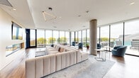 This contemporary amenity space is thoughtfully designed across a single level, offering a variety of distinct zones for relaxation, entertainment, and socializing. The interior is anchored by a sophisticated color palette of slate blue, charcoal, and warm wood tones, carried consistently throughout by rich hardwood flooring and carefully curated artwork.

The main lounge area features expansive floor-to-ceiling glass walls that flood the space with natural light while providing sweeping views of the surrounding landscape and waterfront. A generously sized sectional sofa anchors the seating area alongside a dramatic built-in linear fireplace topped with a large display screen. Decorative open shelving and curated objects add visual interest to the dark accent wall.

Adjacent to the lounge, a dedicated game room includes a full-size pool table, ping-pong table, arcade machine, and shuffleboard, all framed by additional glass walls overlooking a resort-style outdoor terrace and pool. A well-appointed communal kitchen features a large butcher-block dining table, pendant lighting, full cabinetry, and integrated appliances. Throughout, statement light fixtures, gold-accented sconces, and designer furniture contribute to a polished, upscale ambiance.