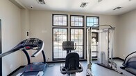 This fitness center is designed to support a comprehensive workout experience within a clean, functional layout spread across a single floor. The main exercise area features durable light-toned flooring and is bathed in natural light through a striking bank of large, black-framed grid windows that overlook mature trees and outdoor surroundings, creating an open and energizing atmosphere. A ceiling fan overhead aids in air circulation throughout the space. The workout floor is outfitted with a variety of cardio machines, including a treadmill, stationary bike, and elliptical, as well as a multi-function cable resistance machine and a weight bench, offering users a well-rounded selection of fitness options. A secondary area, partially separated by a half-wall and reception-style counter, transitions into a dedicated accessory zone. This room houses an assortment of stability and medicine balls displayed on wall-mounted racks, along with additional exercise equipment, making it ideal for stretching, functional training, or group fitness activities. The space is finished with neutral white walls, recessed and surface-mounted lighting, and patterned area rugs that define transitional zones. A keyed exterior door with glass panel provides direct outdoor access, enhancing convenience and ventilation. The overall design is practical, bright, and purposefully organized for an effective fitness experience.