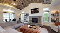This single-floor clubhouse is a well-appointed gathering space that blends rustic charm with modern sophistication. The centerpiece of the main lounge is a striking stacked-stone fireplace framed by shiplap accent walls and flanked by built-in white bookshelves, with a wall-mounted flat-screen television positioned above. A tufted leather ottoman anchors the seating area, which is complemented by a mix of upholstered chairs and sofas arranged for comfortable socializing. Overhead, a reclaimed wood plank ceiling adds warmth and texture, while a geometric chandelier provides an industrial-inspired accent.

Adjacent to the lounge, a quiet seating nook features a pair of elegant arm chairs positioned beside large windows that frame views of the surrounding greenery, inviting natural light throughout the day. High transom windows further enhance the bright, airy atmosphere across the space.

The clubhouse also includes a stylish kitchen outfitted with deep navy cabinetry, brass hardware, white quartz countertops, a subway tile backsplash, and stainless steel appliances, making it fully functional for events and gatherings. Rich dark hardwood flooring runs throughout, tying all areas together cohesively. A casual bar-height dining area with bistro tables and plaid-upholstered stools lines a window wall, offering an inviting spot for informal conversation or light refreshments.