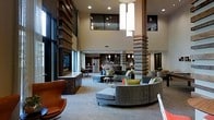 This single-floor communal space makes a striking first impression with its soaring double-height ceilings and an open, airy layout that encourages both relaxation and social gathering. Architectural interest is provided by substantial square columns finished in a combination of wood and stone-like textures, which complement the warm, contemporary design aesthetic throughout. A dramatic pendant light fixture descends from the upper ceiling, serving as a bold focal point and adding to the sense of vertical grandeur.

The main seating area is anchored by a large, curved sectional sofa in soft gray upholstery, paired with accent chairs — including a standout cognac leather piece — and low-profile coffee tables that invite casual conversation. Pops of color from chartreuse and teal throw pillows add vibrancy against the neutral palette. A full-height built-in entertainment wall with a fireplace and a flat-screen TV provides a cozy focal point along one side of the space.

Toward the back, a fully equipped kitchen or kitchenette area is visible, complete with stainless steel appliances and pendant lighting, suggesting a multifunctional space suited for events or resident use. Large windows allow natural light to filter in, further enhancing the open and welcoming atmosphere throughout this thoughtfully designed communal area.