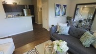 This single-floor residence showcases an open-concept design that integrates the living area and kitchen into one cohesive space. The living room is anchored by a tufted charcoal sofa adorned with layered decorative pillows in neutral and blue-green tones, complemented by a woven coffee table styled with fresh floral arrangements and a geometric patterned area rug. Two framed botanical-style artworks in cool blue and green hues add visual interest to the neutral-toned walls. Rich, dark hardwood-style flooring runs throughout, providing a warm contrast to the light gray walls and creating a sense of continuity across the space. The kitchen, visible beyond a peninsula with light granite countertops, features dark cabinetry and stainless steel appliances, including a refrigerator. A hallway with a paneled white door leads further into the residence, suggesting additional private spaces beyond the main living area. The overall ambiance is calm and well-curated, with a cohesive color palette of grays, greens, and neutrals that lends the space a modern yet inviting character.