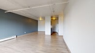 This single-floor unit offers a thoughtfully laid-out living space that balances open-concept design with defined functional areas. The main living area is expansive and open, showcasing light-toned hardwood-style flooring that runs throughout much of the unit. A striking slate-blue accent wall adds depth and character to the space, while exposed conduit and track lighting running along the ceiling lend an industrial aesthetic. Baseboard heating units are positioned along the walls, ensuring year-round comfort. The kitchen is configured as a galley-style layout, lined with dark espresso cabinetry paired with light countertops. It is equipped with a range, refrigerator, dishwasher, and a stainless sink, offering efficient use of space. An adjacent entry area provides two doors and a window, filling the space with natural light and a view of greenery outside. The bedroom features plush carpeting, a window with an outdoor view, and a baseboard heater, creating a warm and comfortable retreat. The bathroom is finished with classic white subway tile surrounding the bathtub and shower, complemented by hardwood-style flooring and a dark vanity. Together, these spaces form a cohesive, modern unit with practical finishes and a clean, contemporary feel.