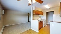 This single-floor residence offers a practical and comfortable open-concept layout, where the living area and kitchen flow naturally into one another. The living space is finished with plush carpeting and neutral-toned walls, creating a warm and inviting atmosphere. A ceiling fan with a light fixture is centrally positioned overhead, providing both air circulation and ambient lighting throughout the space. The kitchen is defined by a half-wall peninsula that subtly separates it from the living area while maintaining an open feel. Rich, warm-toned wood cabinetry lines the upper and lower sections of the kitchen, offering ample storage. A white electric range and oven sit beneath the upper cabinets, accompanied by an overhead ventilation hood. The kitchen floor transitions to hardwood-style flooring, providing a clean contrast to the carpeted living area. A skylight above the kitchen brings in natural light, brightening the workspace. A hallway visible from the main area leads to additional rooms, hinting at a thoughtful and functional floor plan. Overall, the space presents a cozy, well-organized interior with classic residential finishes throughout.