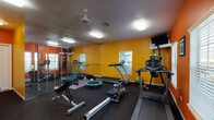 This energetic fitness space is designed to motivate and accommodate a wide range of workout styles. The room's most immediately striking feature is its bold color scheme — vivid orange and yellow walls create an energizing atmosphere, complemented by the motivational wall decal reading "Strong is the New Skinny." Dark rubber flooring covers the entire area, providing durable, slip-resistant support for all types of exercise.

The cardio section is well-stocked, featuring multiple treadmills, an elliptical trainer, and a rowing machine, giving users diverse options for aerobic training. A ceiling-mounted fan aids ventilation and comfort during intense sessions. A wall-mounted flat-screen television is positioned for easy viewing from cardio equipment.

A large mirrored wall on one side of the room visually expands the space and allows users to monitor their form during workouts. A ballet-style barre runs along the mirrored wall, supporting flexibility and balance training. Additional equipment includes a punching bag with stand, a mini trampoline, a weight bench, dumbbells, kettlebells, resistance bands, and a stability ball.

Natural light filters in through multiple windows with white blinds, creating a bright and welcoming environment. The space offers a comprehensive, versatile setup suitable for both cardio and strength training.