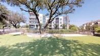 Located at 8233–8241 Meadow Rd in Dallas, Texas, this amenity space combines a well-appointed fitness center with a thoughtfully designed outdoor area. The exterior grounds feature an expansive, well-maintained lawn shaded by mature trees, enclosed by wrought iron fencing and accented with stone seating elements and manicured landscaping. String lights are strung across the outdoor area, adding a welcoming ambiance. The backdrop is a contemporary multi-story residential building with a modern facade of neutral tones, large windows, and private balconies.

Inside, the fitness center is divided into distinct functional zones. The main gym floor is spacious and well-lit with recessed and track lighting, housing a broad array of cardio machines including treadmills, as well as strength training equipment, all set on patterned carpet with yellow accent details. A dedicated studio space features wood-look flooring, floor-to-ceiling mirrors, cycling bikes, a full rack of colorful dumbbells, stability balls, resistance bands, and a BOSU trainer. A large mounted display screen with an interactive fitness platform serves as a focal point in the studio. A ceiling fan and recessed lighting keep the space comfortable. Together, these amenities offer a comprehensive wellness environment suited for a wide range of fitness routines.