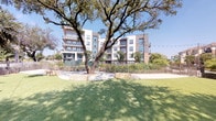 Located at 8233–8241 Meadow Rd in Dallas, Texas, this amenity space combines a well-appointed fitness center with a thoughtfully designed outdoor area. The exterior grounds feature an expansive, well-maintained lawn shaded by mature trees, enclosed by wrought iron fencing and accented with stone seating elements and manicured landscaping. String lights are strung across the outdoor area, adding a welcoming ambiance. The backdrop is a contemporary multi-story residential building with a modern facade of neutral tones, large windows, and private balconies.

Inside, the fitness center is divided into distinct functional zones. The main gym floor is spacious and well-lit with recessed and track lighting, housing a broad array of cardio machines including treadmills, as well as strength training equipment, all set on patterned carpet with yellow accent details. A dedicated studio space features wood-look flooring, floor-to-ceiling mirrors, cycling bikes, a full rack of colorful dumbbells, stability balls, resistance bands, and a BOSU trainer. A large mounted display screen with an interactive fitness platform serves as a focal point in the studio. A ceiling fan and recessed lighting keep the space comfortable. Together, these amenities offer a comprehensive wellness environment suited for a wide range of fitness routines.