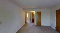 This single-floor residence presents a straightforward and functional layout designed for comfortable everyday living. The interior is finished with neutral wall-to-wall carpeting throughout, providing a soft, cohesive feel across the main living areas. Walls are painted in a clean, bright white, creating a light and open atmosphere that makes the space feel welcoming.

The main living area is open and unobstructed, offering ample room for furnishing and personalization. From this central space, a hallway connects to additional rooms, with warm wood-toned doors providing visual contrast against the white walls and trim. The entry area, visible from the main room, features a narrow window beside the front door that allows natural light to filter into the interior corridor, brightening the transition space.

Multiple doors along the hallway suggest access to private rooms and storage, all fitted with matching medium-toned wooden doors that contribute a consistent, classic aesthetic throughout the home. The ceiling is smooth and flat, maintaining a clean, unadorned look. Overall, the space offers a practical and neutral canvas well-suited for customization, with a simple design that emphasizes functionality and ease of use across its single level.