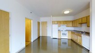 This single-floor unit features an open-concept design that seamlessly connects the living area to the kitchen, creating a spacious and functional layout. The polished concrete-style flooring extends throughout the main space, adding a sleek, modern aesthetic while remaining highly durable and easy to maintain.

The kitchen is positioned along one wall and is outfitted with light wood cabinetry that extends to the upper portion of the walls, providing ample storage. It includes a full suite of white appliances — a refrigerator, electric range with overhead ventilation, and generous counter space with dark laminate countertops. The semi-open partition wall between the kitchen and living area provides slight definition while maintaining an airy, connected feel.

The living area is notably open and unobstructed, with white walls that enhance the sense of brightness in the space. A flush-mount ceiling light fixture provides overhead illumination. Two interior doors visible from the main space suggest access to additional rooms or storage areas. Overall, the unit presents a clean, no-frills environment well-suited for practical, everyday living.